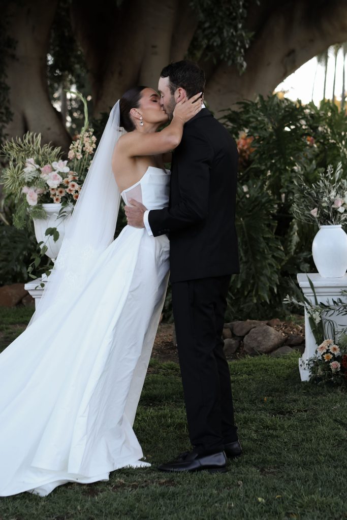 Bride and groom share their first kiss under the Summer House fig trees at Jimbour House, surrounded by floral arrangements and lush garden greenery.
