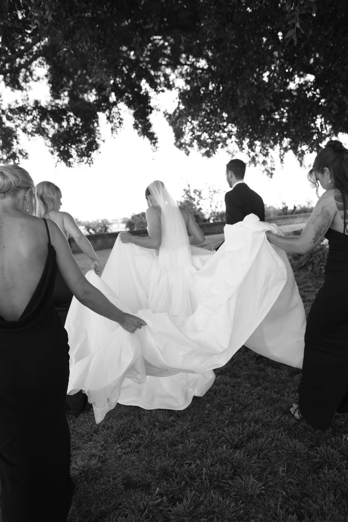 Bride walking through the gardens at Jimbour House with her bridal party helping carry the train of her gown beneath the shade of large fig trees.