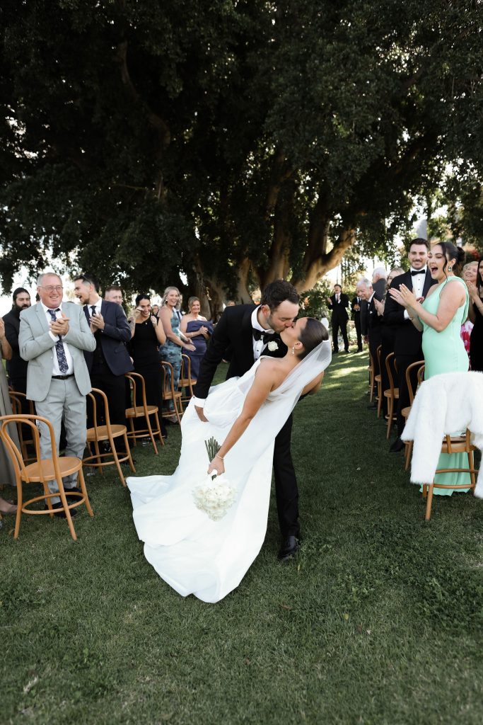 Bride and groom share a kiss during outdoor wedding ceremony recessional at Jimbour House in Queensland, with guests applauding under grand fig trees at this destination wedding venue.