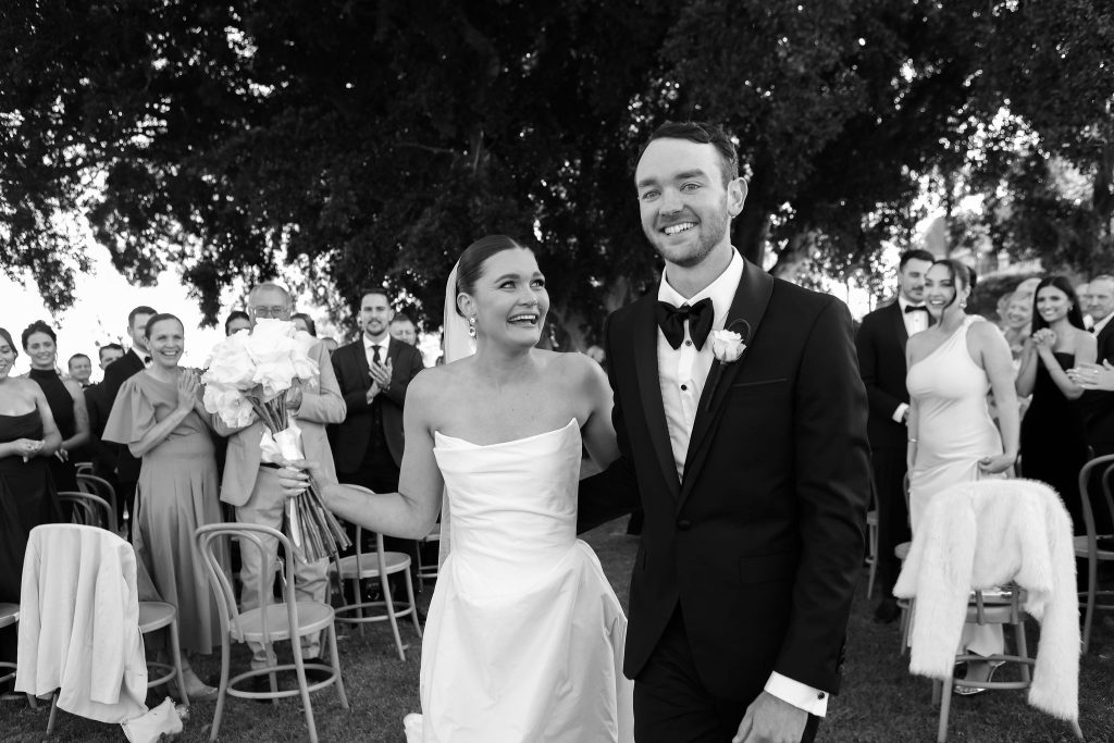 Bride and groom walking up the aisle at Jimbour House surrounded by cheering guests, captured in black and white under the Summer House Fig Trees after their ceremony.