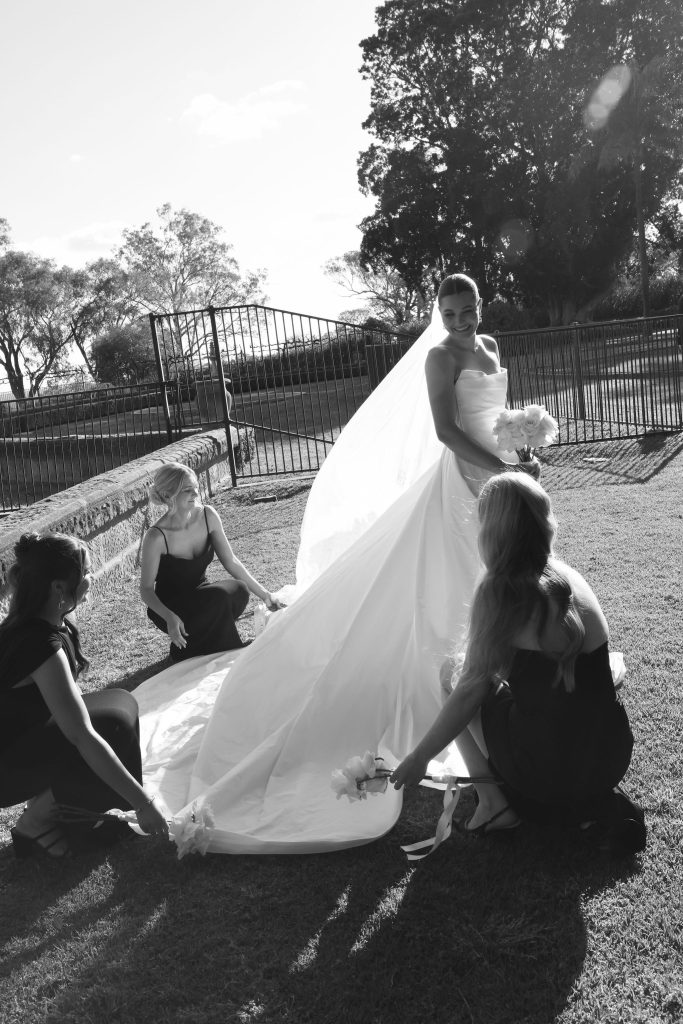 Bride smiling in strapless gown with long train at Jimbour House in Queensland, with bridesmaids adjusting her dress before the outdoor wedding ceremony at this destination venue.