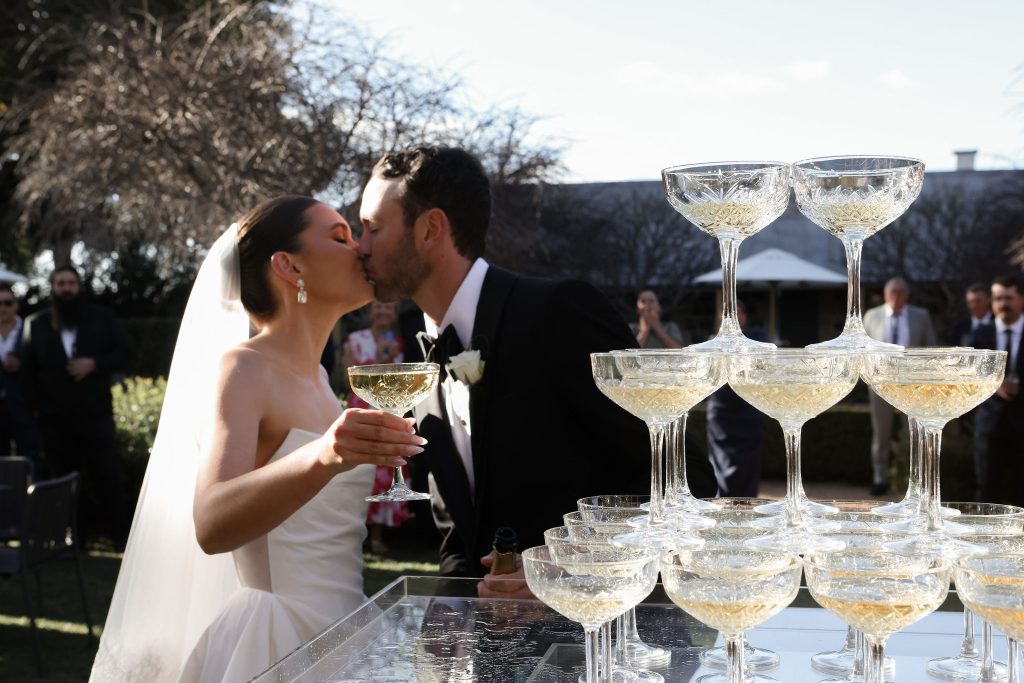 Bride and groom share a kiss during their champagne tower pour at a Jimbour House outdoor wedding reception. The bride holds a crystal coupe filled with champagne while wedding guests look on in the background under soft afternoon light.
