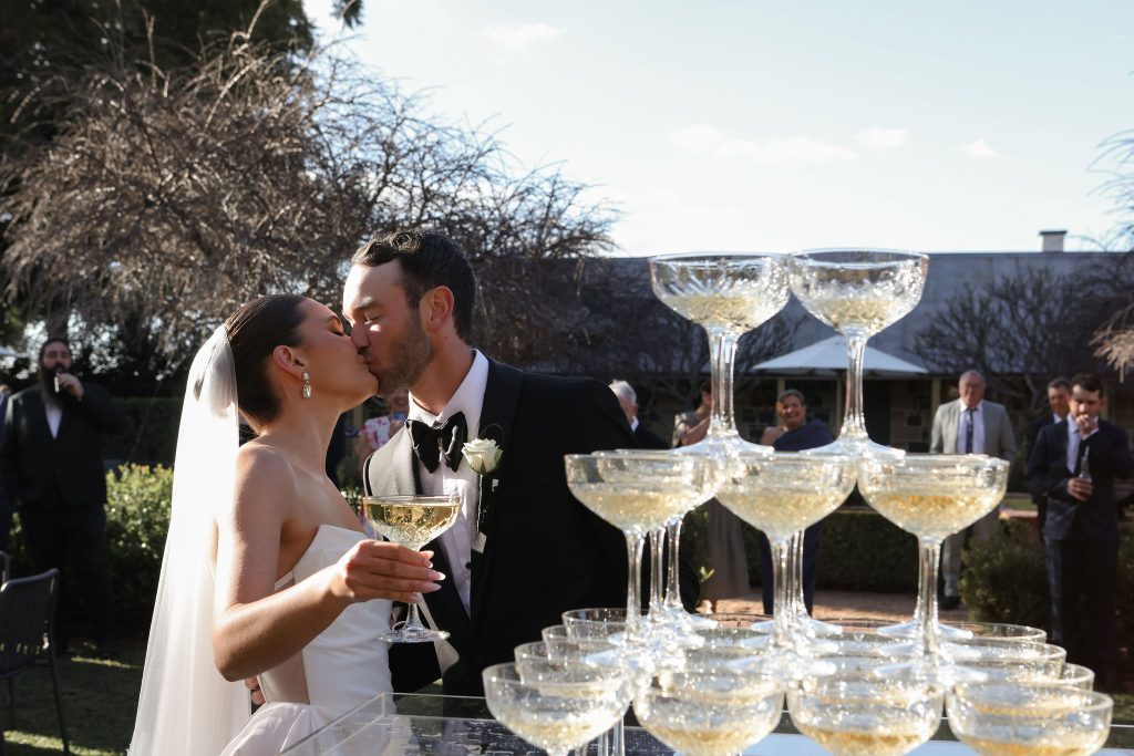 Bride and groom sharing a kiss during outdoor champagne tower celebration at Jimbour House wedding venue in Queensland, a romantic destination for luxury weddings.