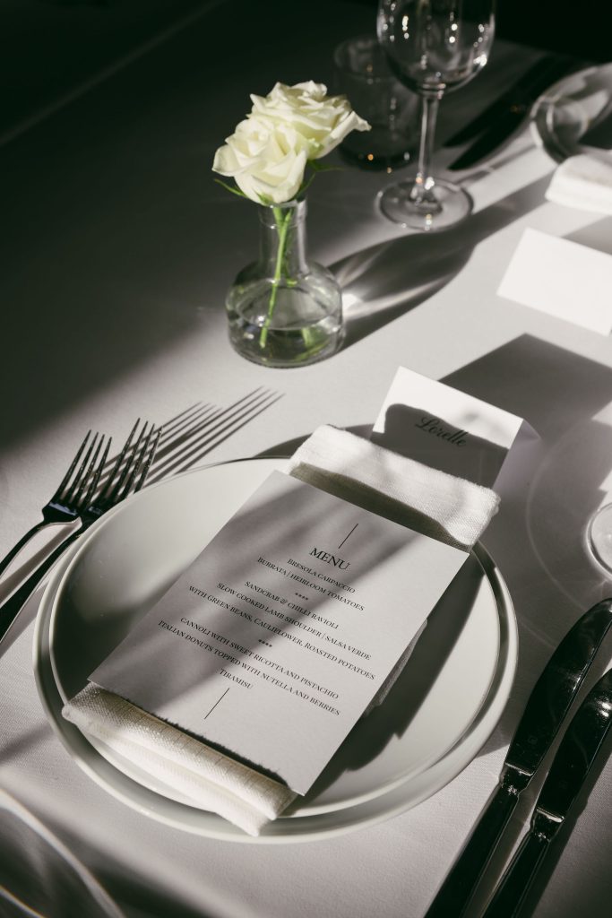 Close-up of a minimalist wedding table setting inside The Hangar at Jimbour House, featuring a printed menu, white linen, and a single white rose in a glass vase.