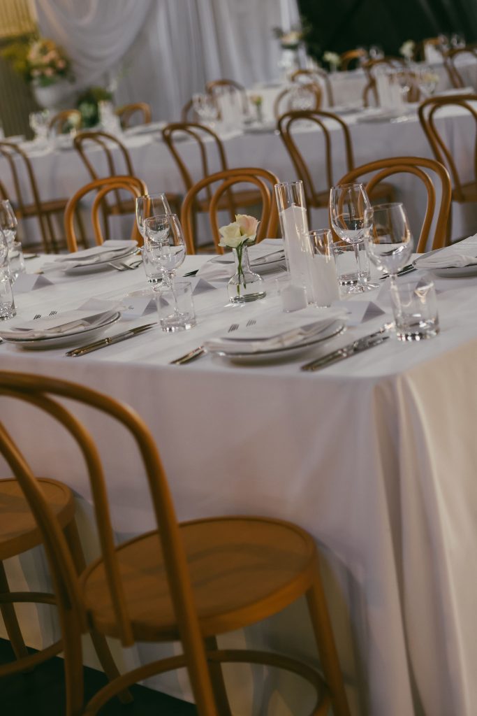 Wedding reception setup inside The Hangar at Jimbour House featuring white tablecloths, glassware, and timber chairs with soft candlelight and minimalist floral details.