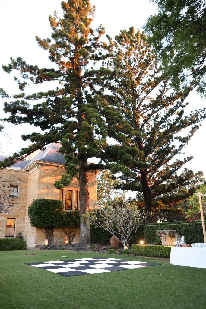 Outdoor lawn area at Jimbour House featuring a black and white checkered dance floor, surrounded by manicured hedges and heritage sandstone walls, framed by tall pine trees in golden hour light