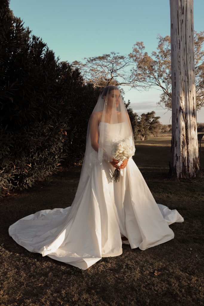 Bride standing in the gardens at Jimbour House wearing a strapless gown and veil, holding a white bouquet in the afternoon light, captured by Kaiti Yann.
