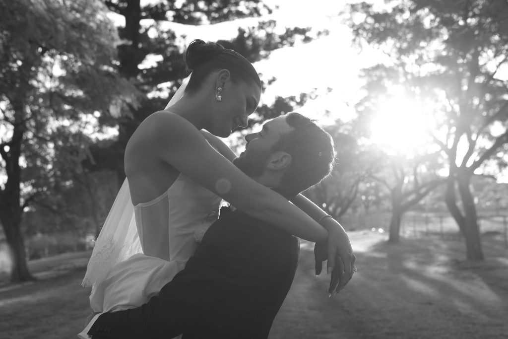 Bride and groom embracing outdoors at Jimbour House with sunlight streaming through the trees, captured in soft black and white by Kaiti Yann.