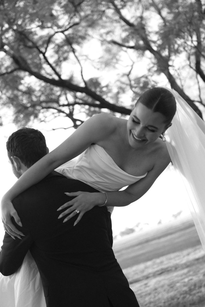 Bride laughing as she’s lifted by the groom in the gardens at Jimbour House, captured in black and white with trees framing the background.