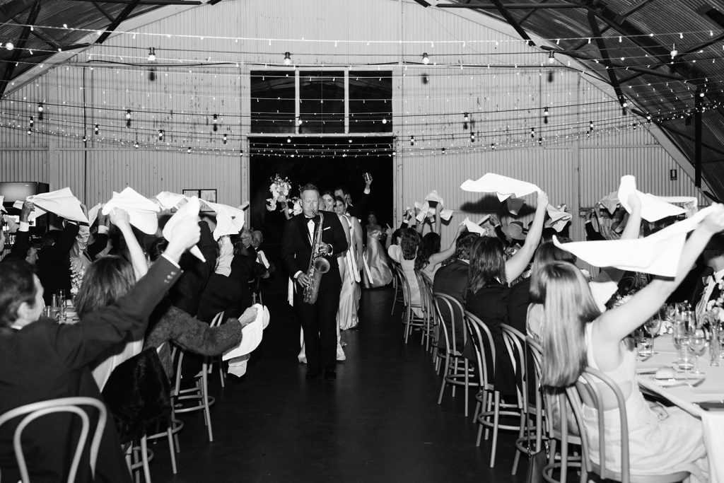 Black and white photo of a wedding party entering The Hangar at Jimbour House with a live saxophonist, as guests wave napkins in celebration under fairy lights.
