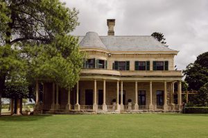 Exterior view of Jimbour House with its sandstone façade, green shutters, and wraparound verandah, framed by lush trees and manicured lawn under a cloudy sky