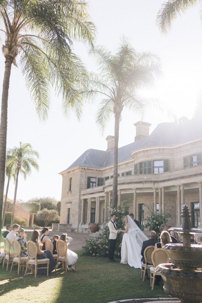  Outdoor garden ceremony at Jimbour House with bride and groom exchanging vows under palm trees in front of the sandstone homestead, surrounded by close family and friends.