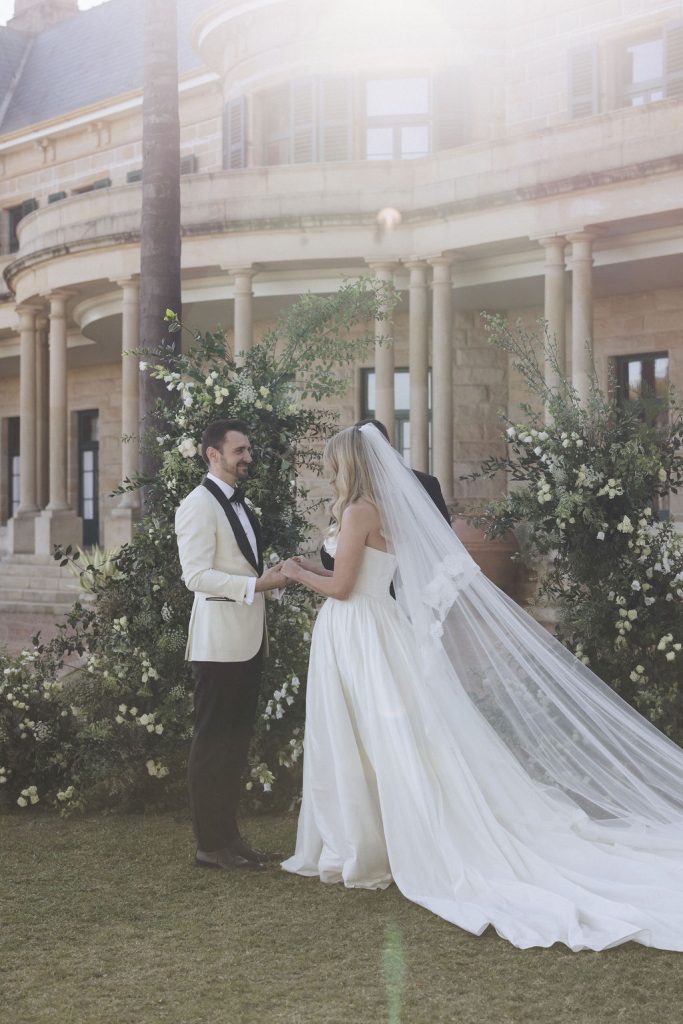Bride and groom exchanging vows during an outdoor ceremony at Jimbour House, framed by lush floral arrangements and the estate’s sandstone facade.