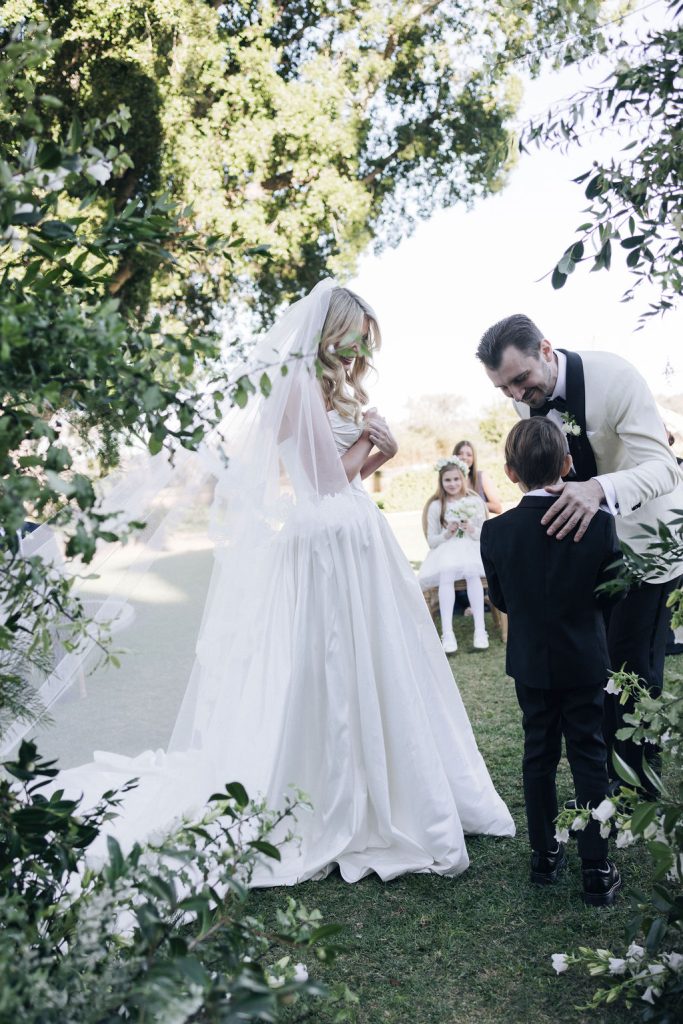 Candid moment between bride, groom, and young page boy during intimate outdoor ceremony at Jimbour House, surrounded by greenery and soft garden florals.