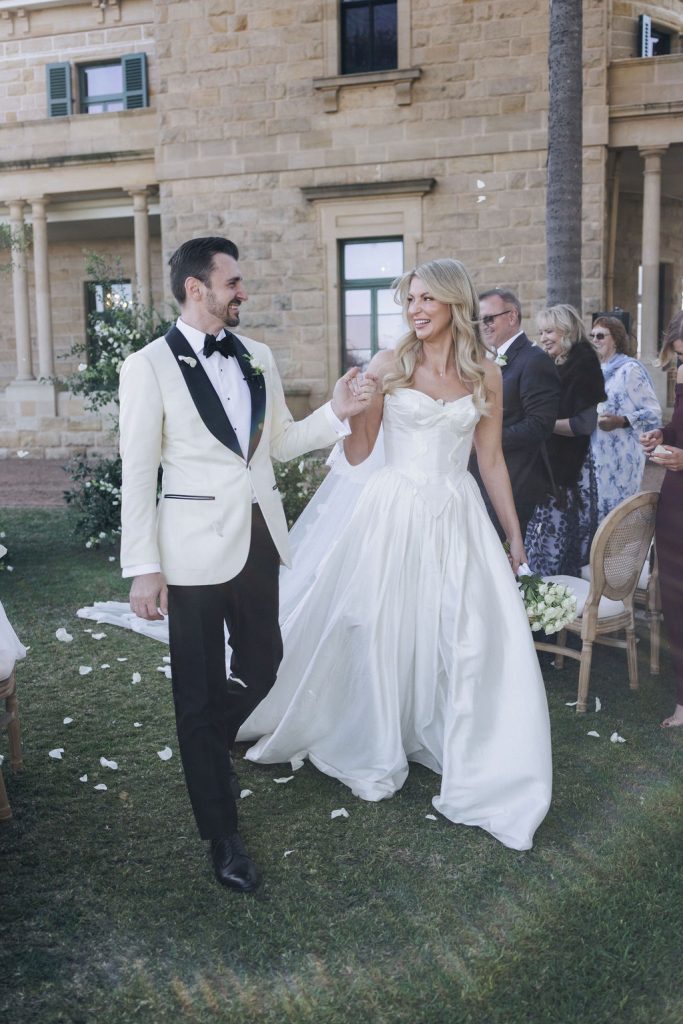 Just married moment at Jimbour House – bride and groom walking hand-in-hand through their ceremony aisle, surrounded by smiling guests and rose petals on the lawn.
