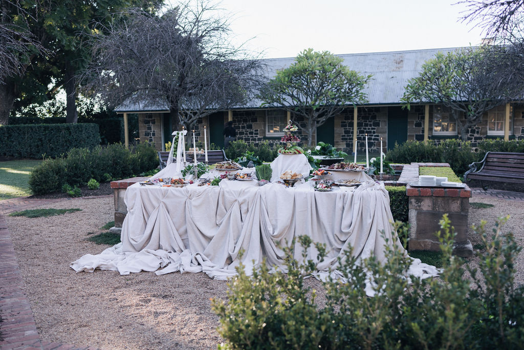 Elegant grazing table styled for a courtyard wedding at Jimbour House, featuring tiered platters, fresh produce, and draped neutral linens against a backdrop of heritage buildings and manicured gardens.