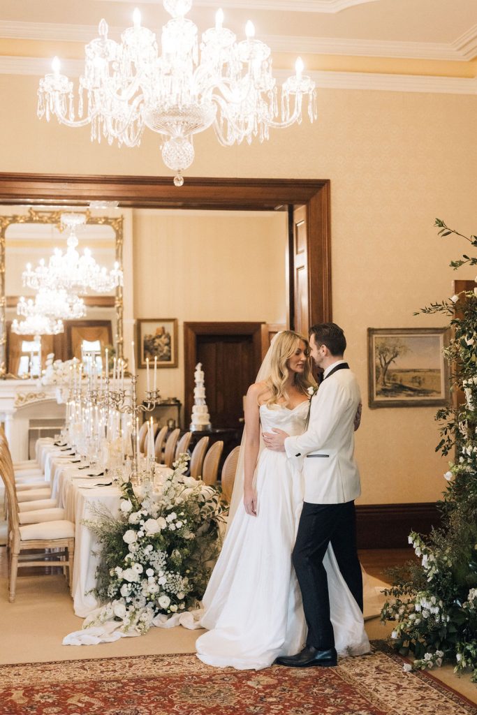 Bride and groom share a quiet moment in the formal dining room of Jimbour House, surrounded by chandeliers, grand floral arrangements, and a long candlelit table set for their elegant reception. A tiered wedding cake stands in the background.