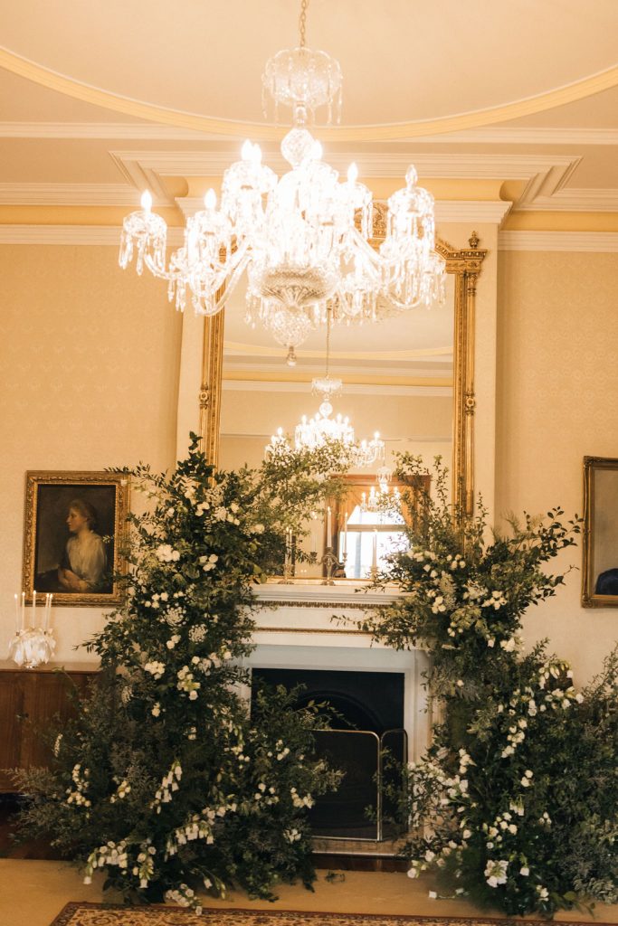 Indoor wedding ceremony setup at Jimbour House featuring lush floral arrangements around the fireplace beneath crystal chandeliers.