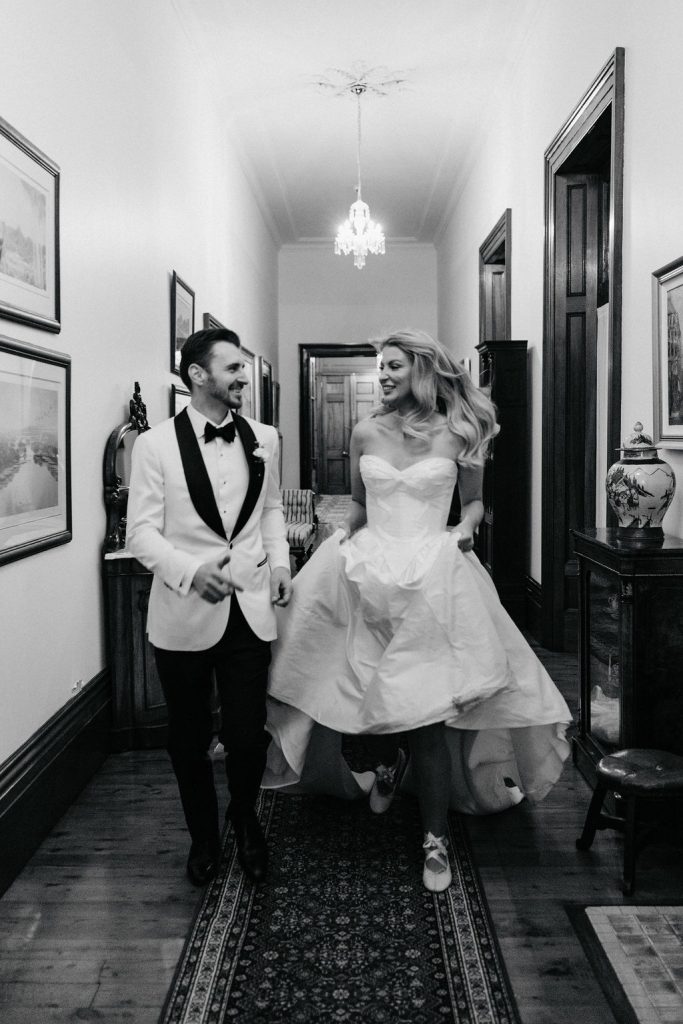 Black and white photo of a joyful bride and groom walking down a hallway inside Jimbour House. The bride lifts her gown to reveal ballet flats, adding a fun, relaxed touch to their formal attire as they smile at each other.