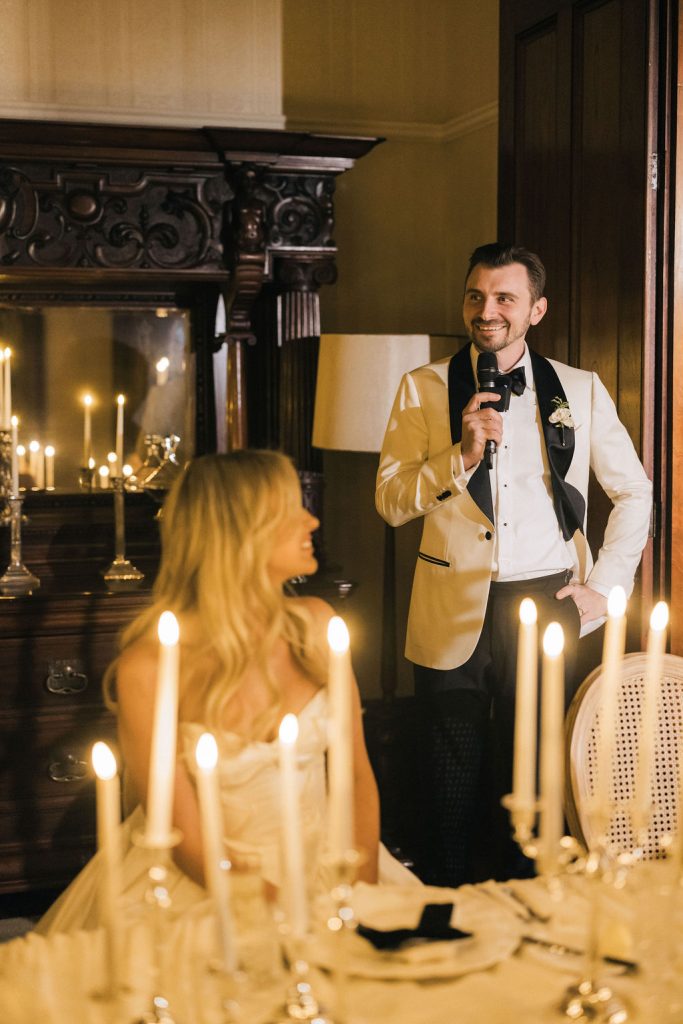 Groom giving a heartfelt wedding speech by candlelight inside Jimbour House, wearing a cream tuxedo jacket, as the bride looks on lovingly from the dinner table surrounded by elegant taper candles.