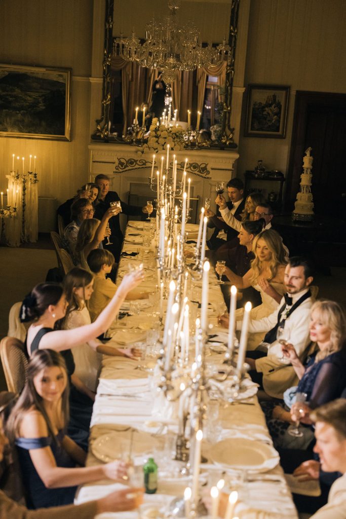 Guests raise their glasses for a candlelit toast during a formal wedding dinner at Jimbour House. The long banquet table is elegantly styled with crystal candelabras and white linen, creating a warm and intimate atmosphere under a grand chandelier.