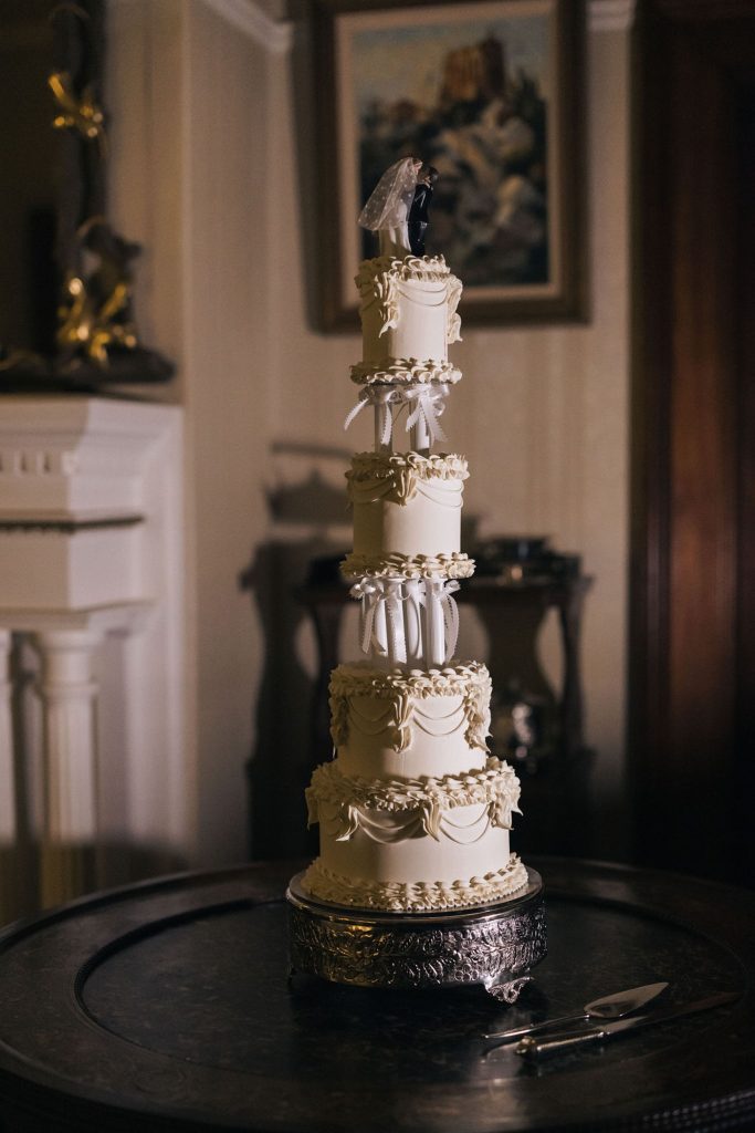 A tall, vintage-inspired wedding cake with seven tiers, piped detailing, and white ribbon accents, topped with a classic bride and groom figurine. The cake sits on an ornate silver stand inside one of Jimbour House’s formal reception rooms.