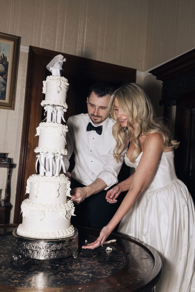  Bride and groom cut into their towering white wedding cake adorned with intricate piping and a vintage-style cake topper. The couple smiles as they share this classic wedding tradition inside one of Jimbour House’s heritage rooms.