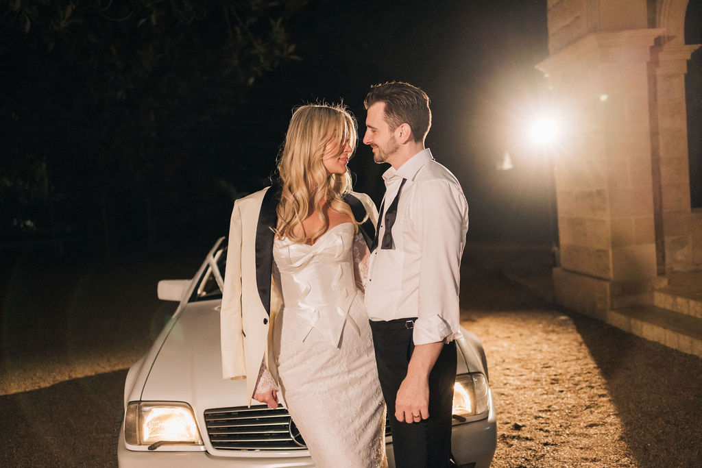 A bride and groom share an intimate moment outside Jimbour House at night, standing in front of a vintage white car with headlights on. The bride wears a sleek reception dress with the groom’s jacket draped over her shoulders, while the groom has his shirt slightly unbuttoned and bowtie undone, capturing the relaxed elegance of their evening celebration.