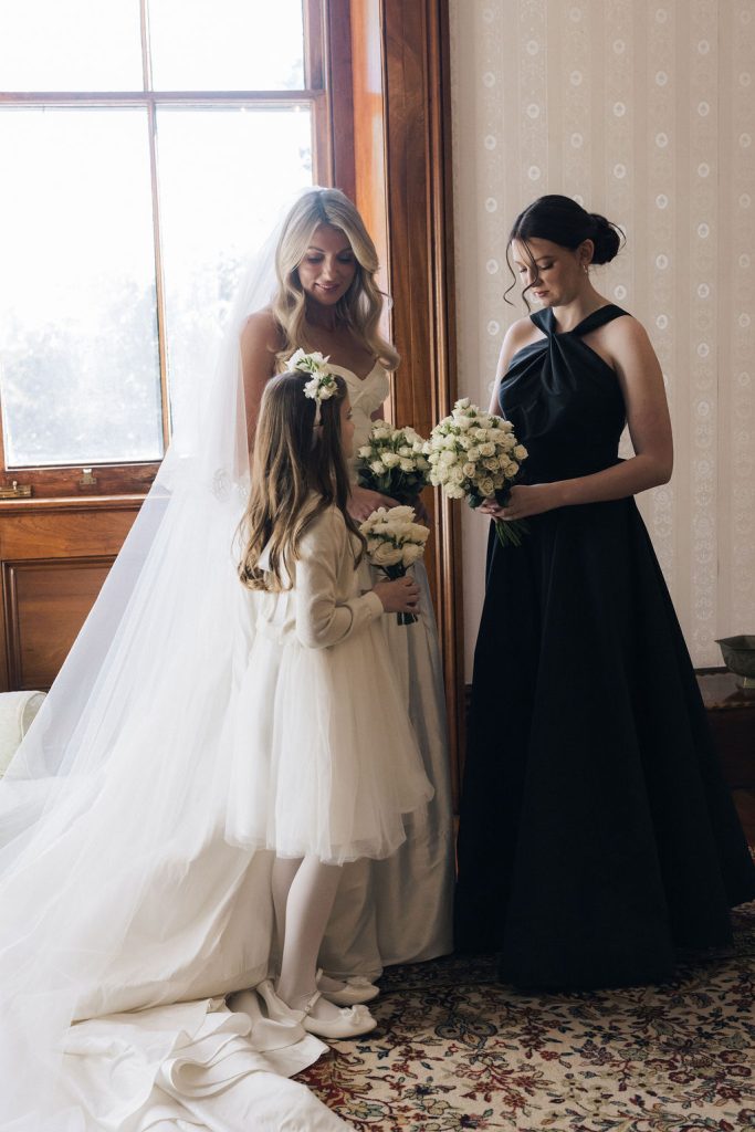 Bride in strapless gown standing with flower girl and bridesmaid, holding white rose bouquets inside a heritage room at Jimbour House before the wedding ceremony.