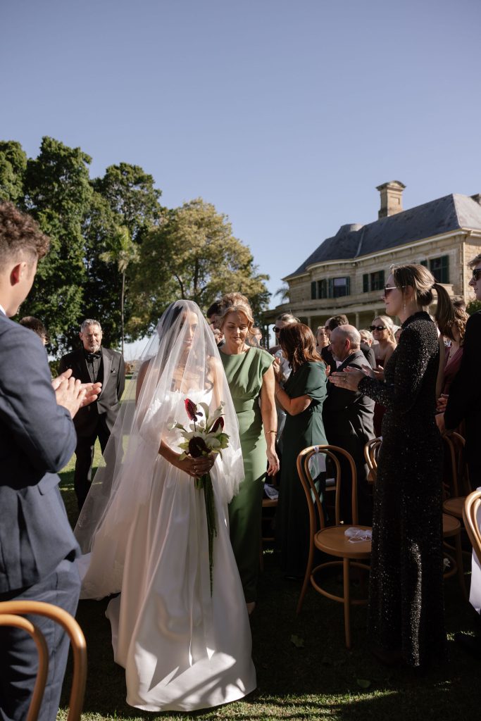 Bride walking down the aisle with veil and bouquet during an outdoor wedding ceremony at Jimbour House, with guests standing and sandstone homestead in the background
