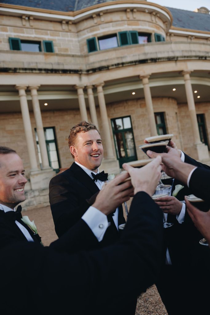 Groom and groomsmen toasting espresso martinis during cocktail hour in front of the sandstone façade at Jimbour House Queensland