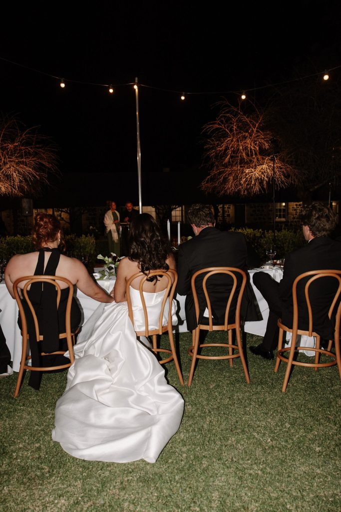Bride seated at outdoor wedding reception under festoon lights during evening speeches at Jimbour House Queensland