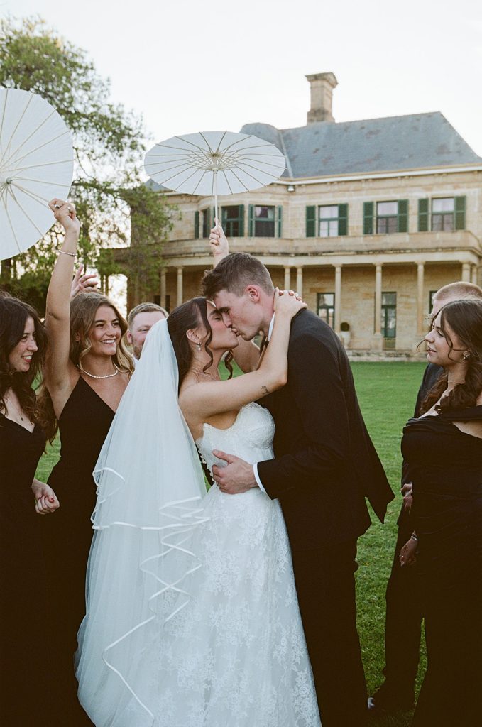Bride and groom kissing on the lawn at Jimbour House with bridal party holding parasols, sandstone homestead in the background at a Queensland wedding venue
