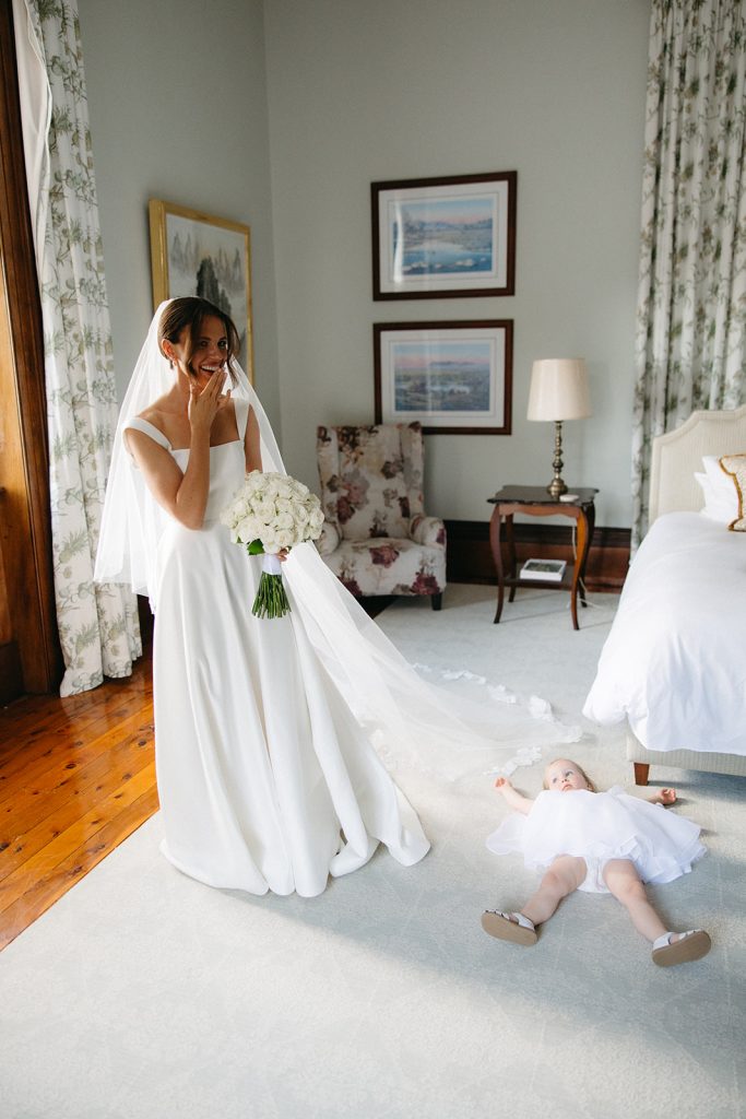Bride in structured white gown holding a rose bouquet inside Jimbour House, with a flower girl lying on the floor in a matching white dress, capturing a candid and playful pre ceremony moment
