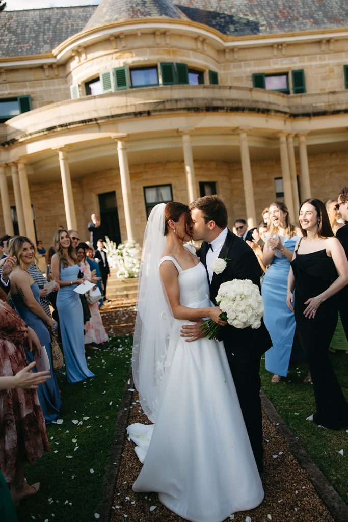 Bride and groom share a kiss after the ceremony at Jimbour House, surrounded by guests on the garden aisle with white petals underfoot and the sandstone homestead behind

