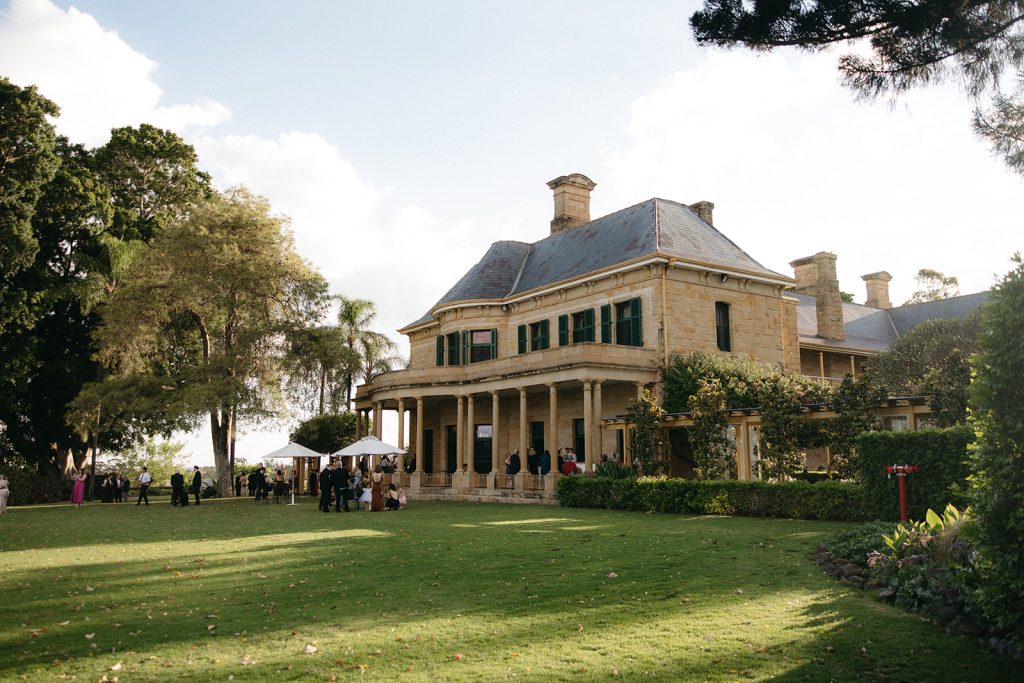 Guests gathering on the lawn at Jimbour House during cocktail hour, with the sandstone homestead and wraparound veranda overlooking the garden setting
