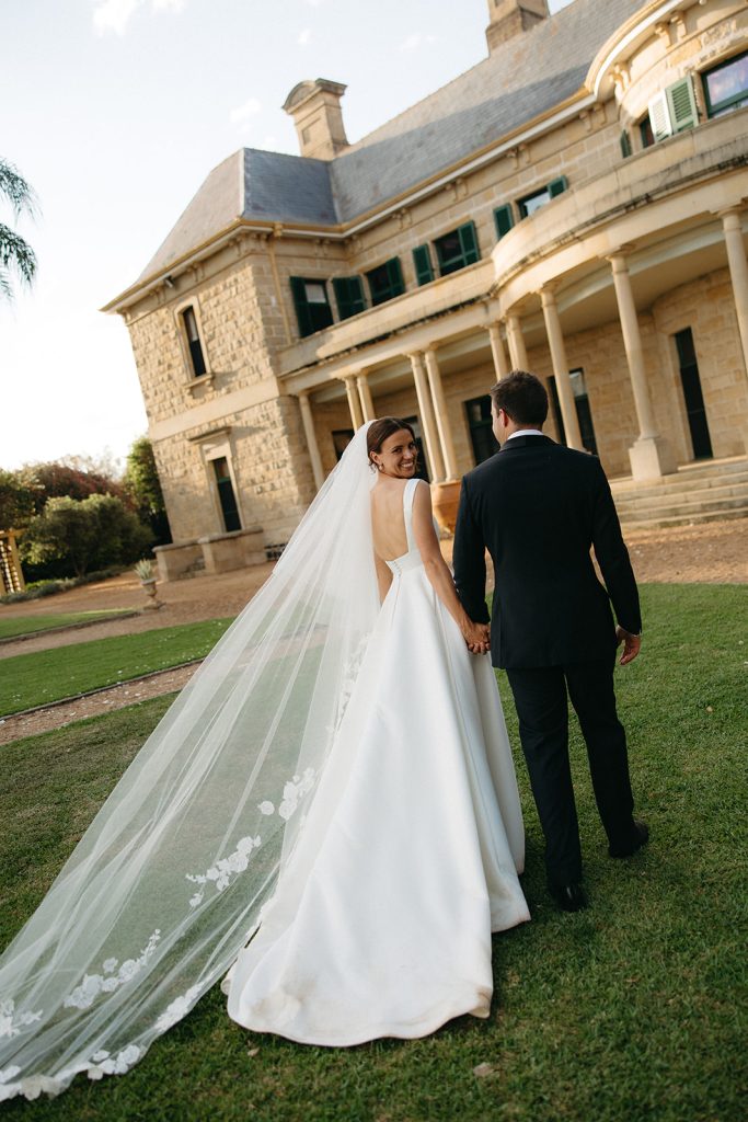 Bride and groom walking across the lawn at Jimbour House, long veil trailing behind, sandstone homestead creating a strong architectural backdrop
