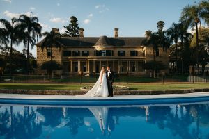 Bride and groom in black tie standing in front of Jimbour House with reflection in the pool, showcasing the grand sandstone homestead and formal garden setting in Queensland