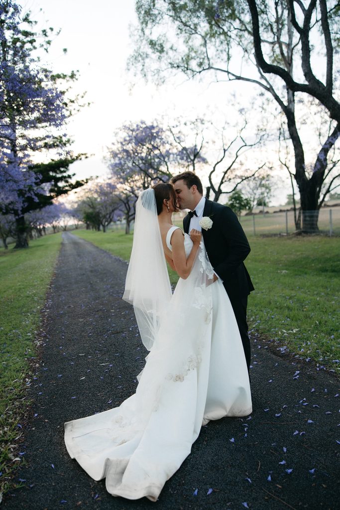 Bride and groom share a quiet kiss along a tree lined driveway at Jimbour House, jacaranda petals scattered across the ground and soft evening light filtering through the branches
