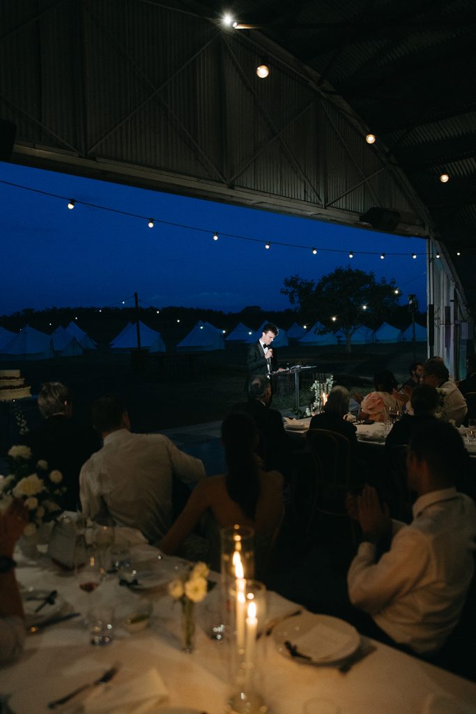 Evening speeches inside the Hangar at Jimbour House, candlelit tables in the foreground and open doors revealing glamping tents under the night sky
