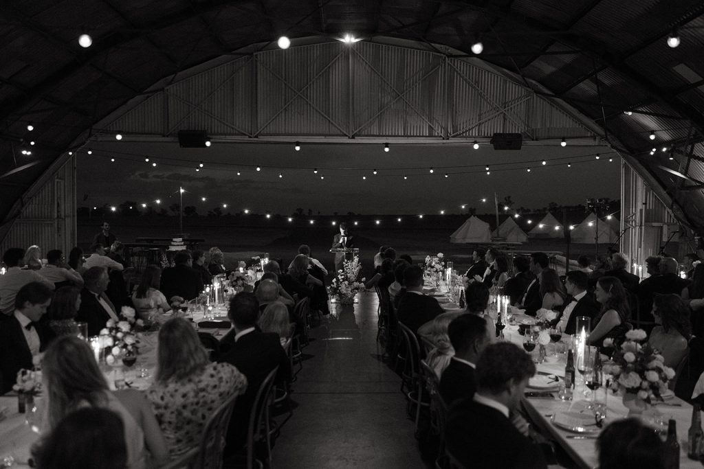 Wide view of the reception inside the Hangar at Jimbour House, long tables set under festoon lighting with guests gathered for speeches and open views to the glamping tents outside
