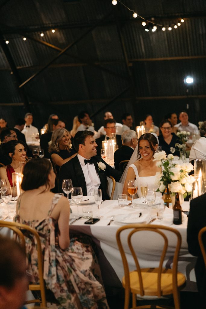 Bride and groom seated among guests during the reception in the Hangar at Jimbour House, candlelit tables and relaxed expressions capturing the atmosphere of the evening
