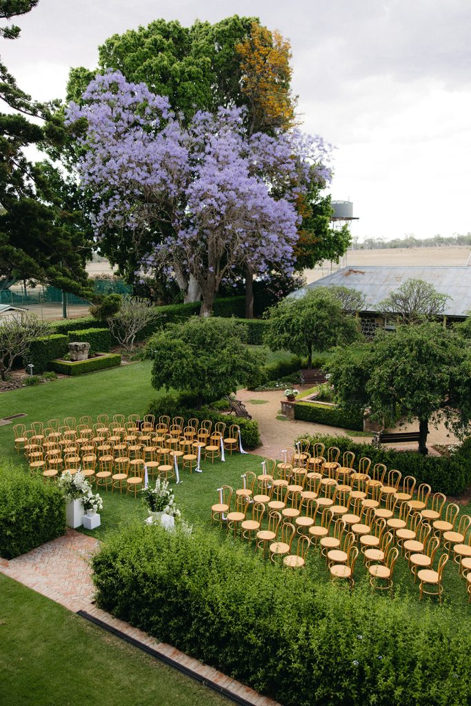 Courtyard ceremony setup at Jimbour House with curved rows of timber chairs, white floral arrangements and manicured hedges, framed by blooming jacaranda trees and garden pathways
