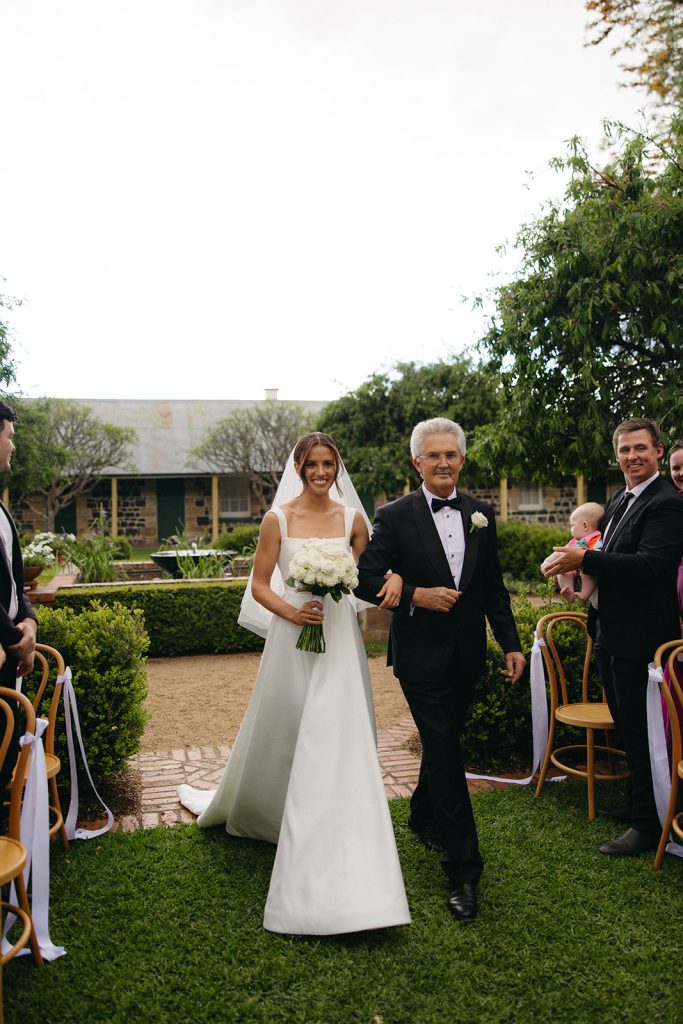 Bride walking down the aisle in the Jimbour House courtyard with her father, holding a white rose bouquet, surrounded by seated guests and garden hedging
