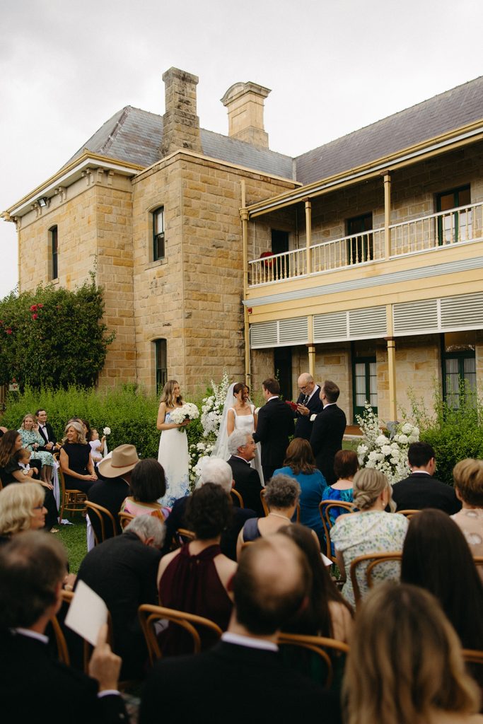 Courtyard wedding ceremony at Jimbour House with bride and groom exchanging vows, surrounded by guests seated on timber chairs and white floral arrangements against the sandstone homestead backdrop
