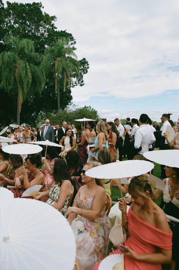 Wedding guests seated and standing on the garden lawn holding parasols during an outdoor ceremony at Jimbour House Queensland, surrounded by palms and greenery
