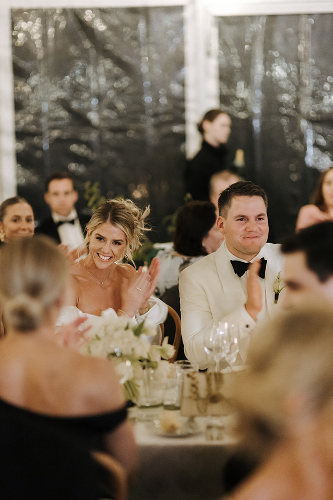 Bride and groom smiling and clapping during their wedding reception inside a clear marquee at Jimbour House.