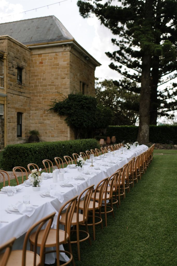  Long outdoor wedding reception table with white linens and timber chairs set beside the sandstone homestead at Jimbour House Queensland, styled on the garden lawn
