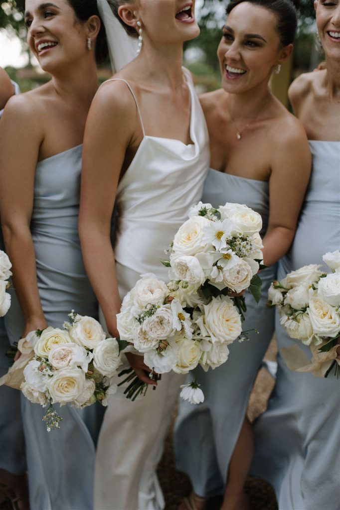 Bride with bridesmaids holding white rose bouquets, wearing satin dresses in soft tones during a garden wedding at Jimbour House Queensland
