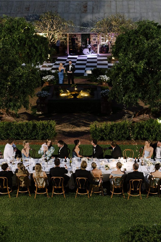Outdoor wedding reception at Jimbour House with long banquet table, guests seated on the lawn, and speeches taking place beside the courtyard fountain under festoon lighting in Queensland
